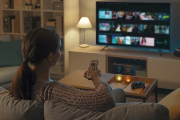 Перегляд сумнівного контенту на Amazon Fire Stick став ще складнішим, оскільки VPN ще не доступні на цьому пристрої. Жінка відпочиває на дивані, користується пультом дистанційного керування та вибирає телешоу або фільм у меню телевізора; Shutterstock ID 2126878775; замовлення_на_продаж: -; робота: -; клієнт: -; інше: -