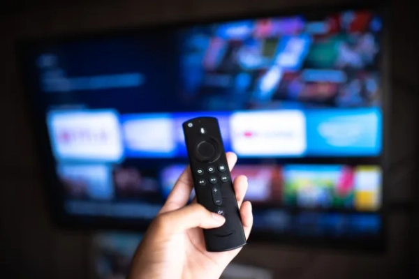 Перегляд сумнівного контенту на Amazon Fire Stick став ще складнішим, оскільки VPN ще не доступні на цьому пристрої. Пульт дистанційного керування Amazon Fire TV Stick у руці з вибірковим фокусом на телевізійному фоні.