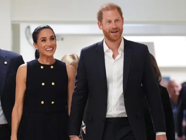 Meghan Markle and Prince Harry walk hand in hand through an indoor corridor in formal black outfits.