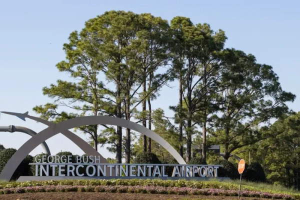 George Bush Intercontinental Airport entrance sign stands beside trees, landscaping, and a stop sign under a clear sky.