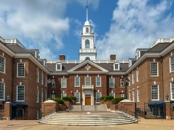 Delaware Legislative Hall’s red brick facade and white cupola stand above broad front steps under a partly cloudy sky.