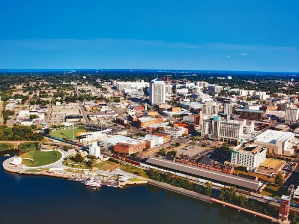 Aerial view of downtown Montgomery beside the Alabama River, with skyline, rail tracks, riverfront park, and baseball stadium.