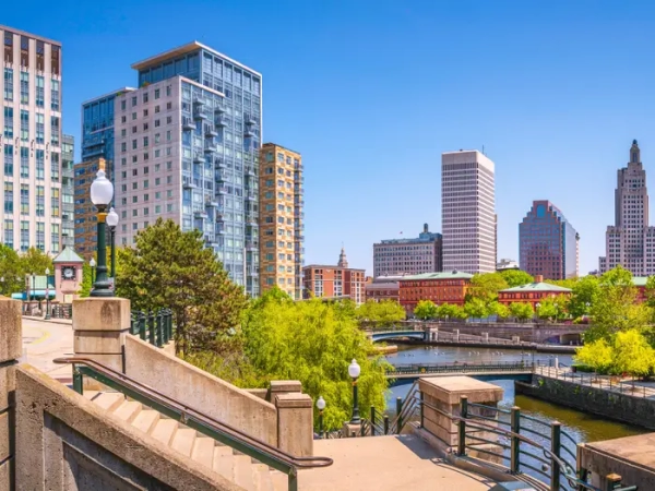 Downtown Providence skyline rises above riverwalk stairs, bridges, trees, and waterfront on a clear sunny day.