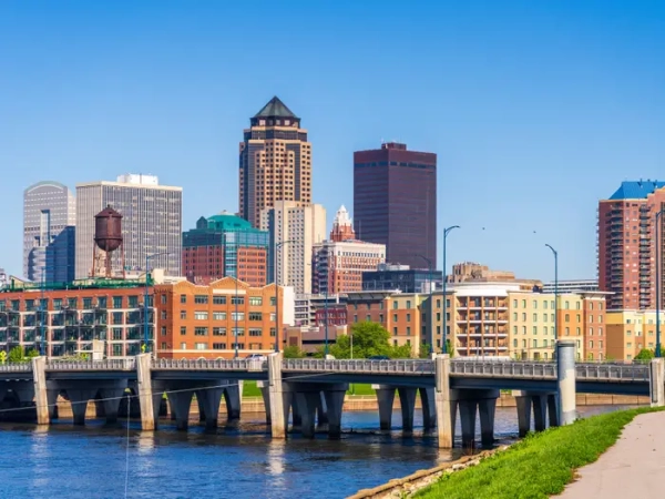 Downtown Des Moines skyline rises behind a bridge over the river on a clear sunny day.