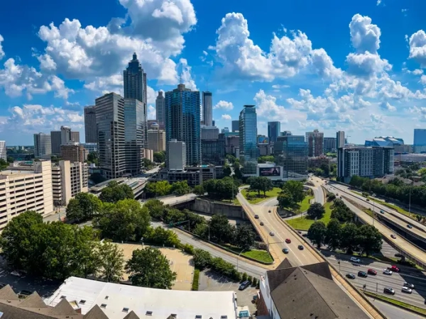 Downtown Atlanta skyline rises above highways and trees under a bright blue sky with large cumulus clouds.