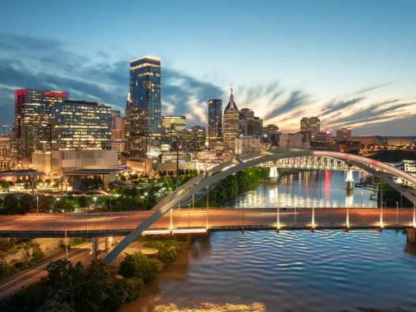 Downtown Nashville skyline glows at dusk behind an arched bridge over the Cumberland River.