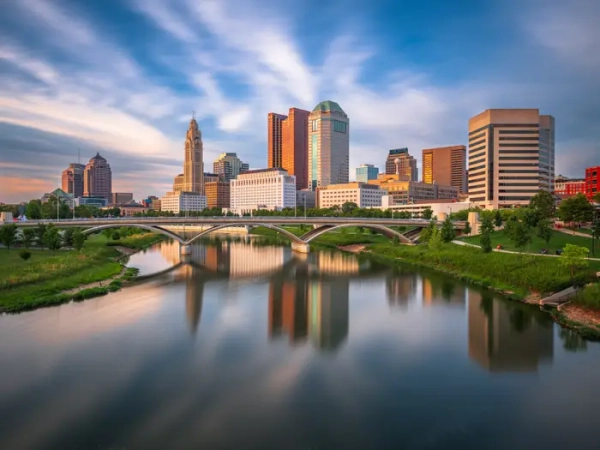 Downtown Columbus skyline and arched bridge reflect in the Scioto River under a streaked evening sky.