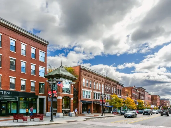 Brick storefronts line a downtown street with cars, American flags, benches, and dramatic cloudy sky.