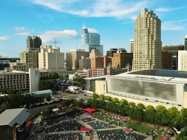 Downtown Raleigh skyline rises behind an outdoor amphitheater and convention center under a blue sky.