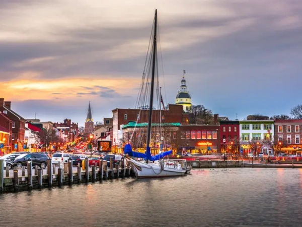 A sailboat moored at Annapolis harbor with downtown streets and the Maryland State House at sunset.