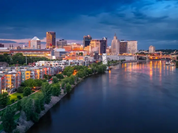 Downtown Saint Paul skyline glows at dusk beside the Mississippi River under a cloudy blue sky.