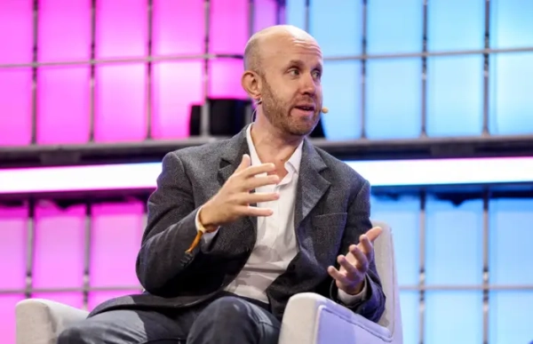 Bald speaker in a blazer gestures while seated onstage before a pink and blue illuminated backdrop.