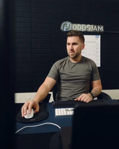 Людина сидить за столом, користуючись мишкою, в офісі зі знаком ODDSJAM на стіні.